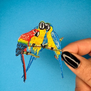 May include: Three embroidered parrots on a branch. The parrots are yellow, blue, and red. The parrots are facing each other.
