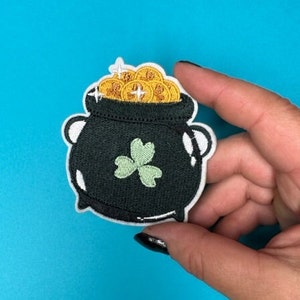 以下が含まれることがあります： 緑のクローバーと金貨が上からこぼれ落ちている黒い鍋。鍋は白い輪郭で囲まれています。