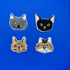 May include: Four embroidered cat face patches. The patches are on a blue background. The cats have different fur colours and patterns.