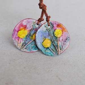 Puede incluir: Dos colgantes circulares de cerámica suspendidos de un cordón marrón. Cada colgante presenta una superficie texturizada con un diseño estilo acuarela en tonos azul, morado y rosa, acentuado por formas florales amarillas en relieve y tallos finos.