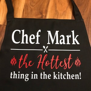 May include: Black apron with white text that reads "Chef Mark" above "the Hottest thing in the kitchen!" in red script. The design includes crossed utensils and flame graphics.