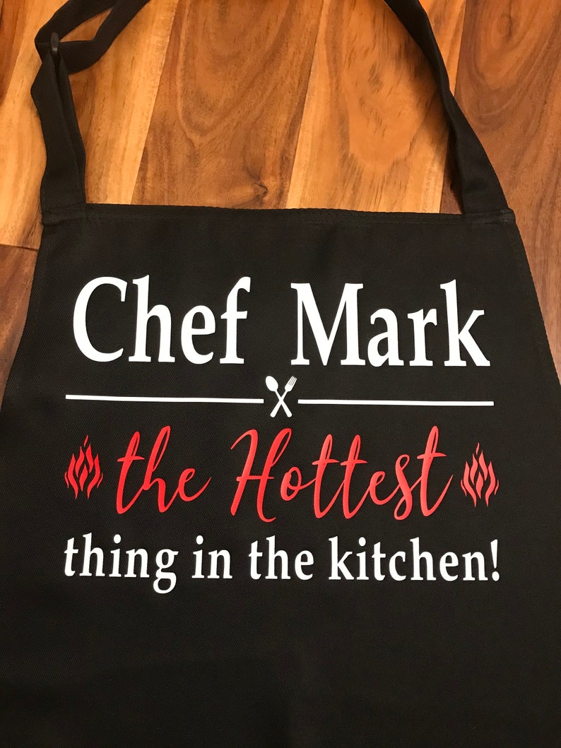 May include: Black apron with white text that reads "Chef Mark" above "the Hottest thing in the kitchen!" in red script. The design includes crossed utensils and flame graphics.