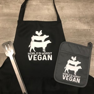 Peut inclure: Tablier de cuisine noir avec un graphique blanc d'un coq, d'un cochon et d'une vache empil&eacute;s les uns sur les autres. Le texte "World's Worst Vegan" est imprim&eacute; sous le graphique. Un gant de four gris assorti avec le m&ecirc;me graphique et le m&ecirc;me texte est inclus.