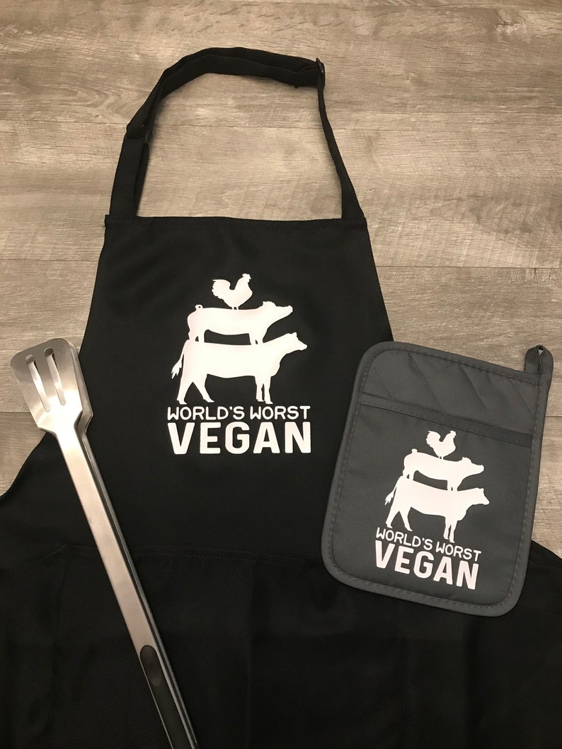 Peut inclure: Tablier de cuisine noir avec un graphique blanc d'un coq, d'un cochon et d'une vache empil&eacute;s les uns sur les autres. Le texte "World's Worst Vegan" est imprim&eacute; sous le graphique. Un gant de four gris assorti avec le m&ecirc;me graphique et le m&ecirc;me texte est inclus.