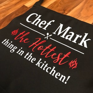 May include: Black apron with the text "Chef Mark the Hottest thing in the kitchen!" in white and red. The apron has a neck strap and waist ties. The text includes a flame graphic.