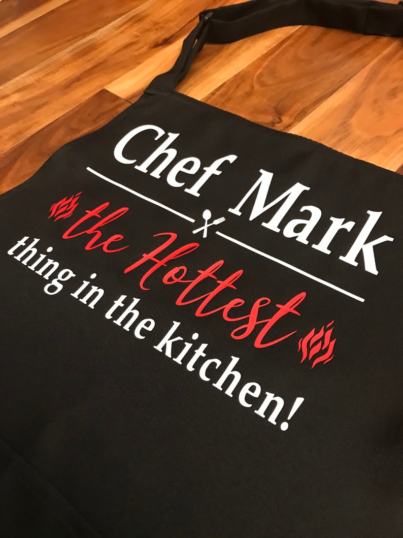 May include: Black apron with the text "Chef Mark the Hottest thing in the kitchen!" in white and red. The apron has a neck strap and waist ties. The text includes a flame graphic.