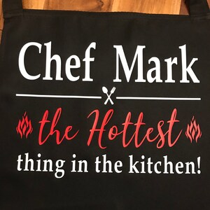 May include: Black apron with the text "Chef Mark" in white, above "the Hottest thing in the kitchen!" in red script. A white line with a spoon and fork graphic separates the phrases. Flames are on either side of "the Hottest".