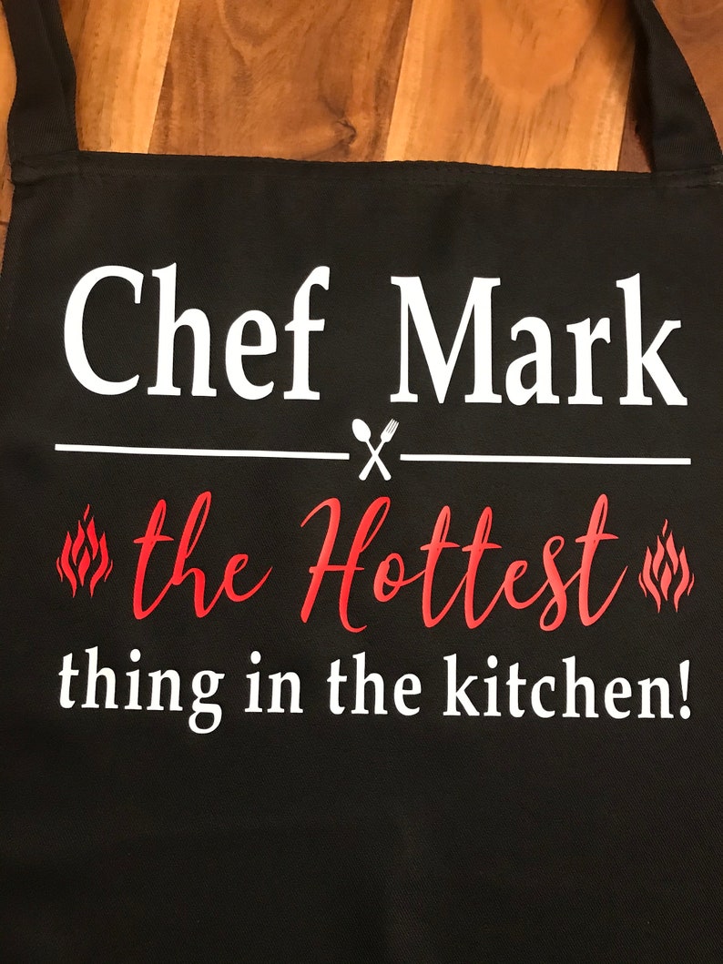 May include: Black apron with the text "Chef Mark" in white, above "the Hottest thing in the kitchen!" in red script. A white line with a spoon and fork graphic separates the phrases. Flames are on either side of "the Hottest".