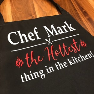 May include: Black apron with the text "Chef Mark" above "the Hottest thing in the kitchen!" in white and red lettering. The apron has a neck strap and is made of a durable fabric.