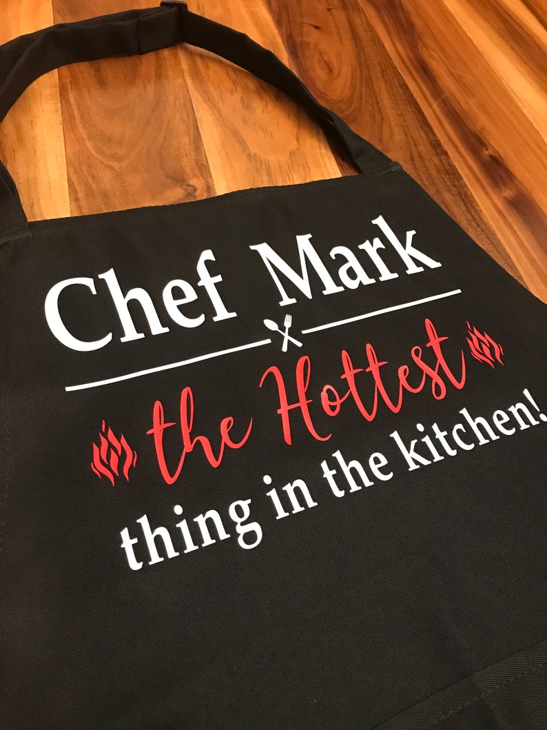 May include: Black apron with the text "Chef Mark" above "the Hottest thing in the kitchen!" in white and red lettering. The apron has a neck strap and is made of a durable fabric.