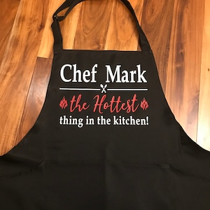 May include: Black apron with the text "Chef Mark" in white, with a crossed knife and fork design. Below, in red script, it reads "the Hottest thing in the kitchen!" The apron has a pocket and adjustable neck strap.