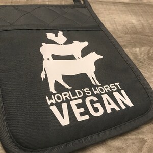 Peut inclure: Un gant de cuisine gris fonc&eacute; avec une silhouette blanche d'un poulet, d'un cochon et d'une vache empil&eacute;s les uns sur les autres. Le texte "WORLD'S WORST VEGAN" est imprim&eacute; en blanc sous les animaux.