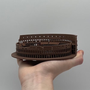 May include: A brown plastic model of the Colosseum, a famous Roman amphitheatre, held in a person's hand.