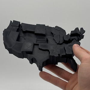 May include: A black, three-dimensional sculpture of the United States, with each state represented as a separate block. The sculpture is held in a hand against a white background. The piece appears to be made of a solid, matte material.