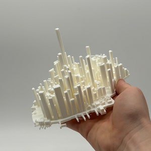 Op de afbeelding: Een 3D-geprint architectuurmodel in gebroken wit, vastgehouden in een hand. Het model toont een stadsgezicht met verschillende rechthoekige en cilindrische structuren van verschillende hoogtes, die lijken op wolkenkrabbers en gebouwen. De achtergrond is effen wit.