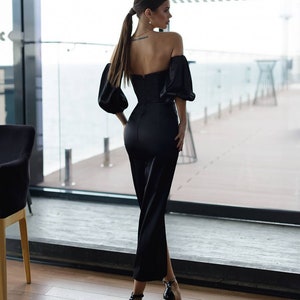 May include: A woman wearing a black off-the-shoulder dress with a fitted bodice and a flowing skirt. She is standing in front of a window with a view of the ocean.
