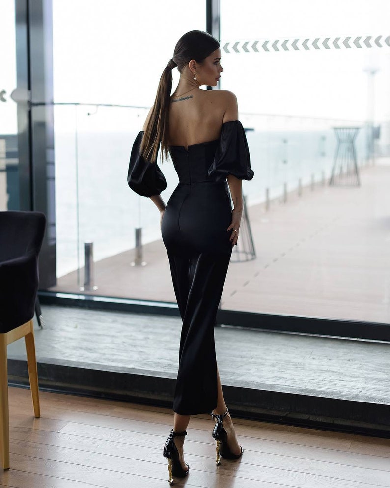 May include: A woman wearing a black off-the-shoulder dress with a fitted bodice and a flowing skirt. She is standing in front of a window with a view of the ocean.