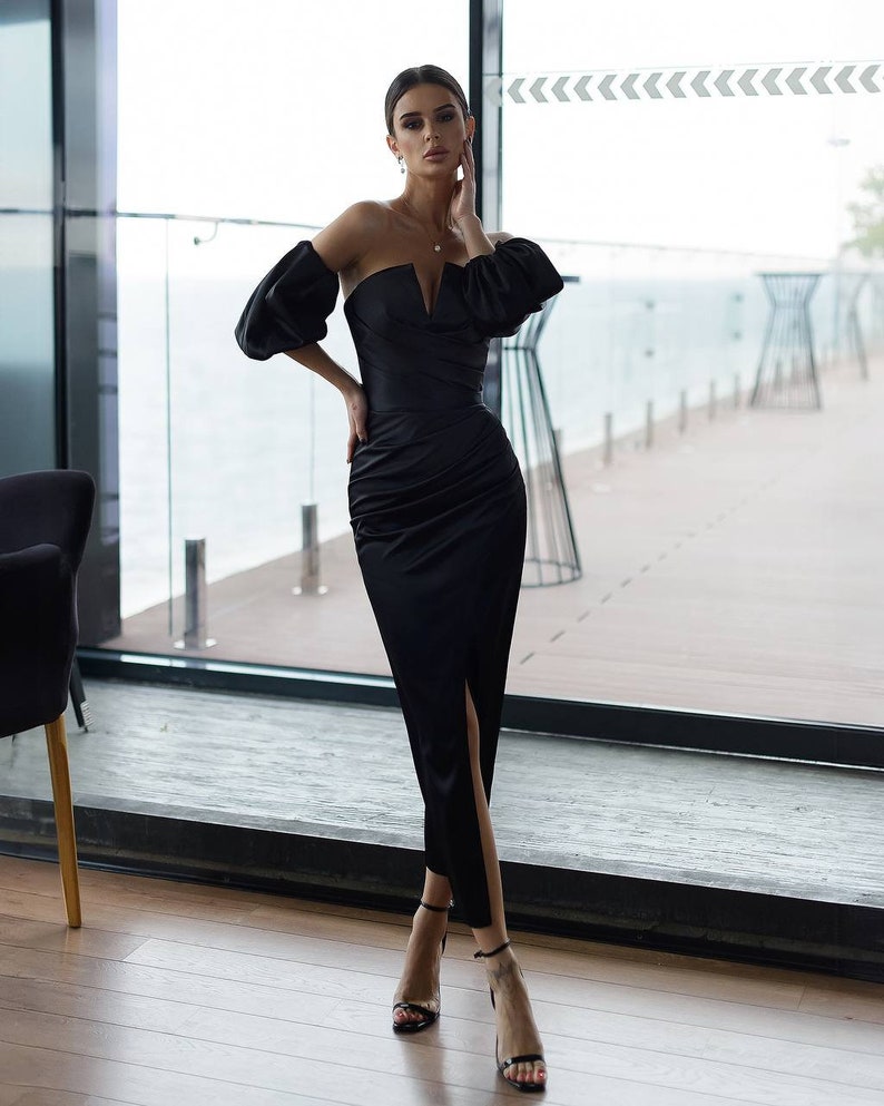 May include: A woman wearing a black strapless dress with a fitted bodice and a thigh-high slit. The dress has puffed sleeves and a V-neckline. She is wearing black heels.