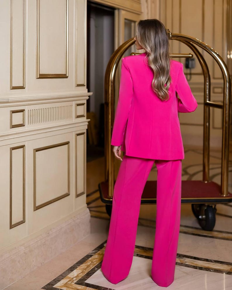 May include: A bright pink pantsuit, featuring a long blazer with shoulder pads and wide-leg trousers. The suit is a vibrant fuchsia color, and the person is standing in a well-lit hallway with decorative wall panels and a gold luggage cart.