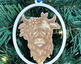 May include: A wooden Christmas ornament featuring a laser-cut design of a Highland cow with a floral crown. The ornament is round and has a white background with a brown cow inside. The ornament is hanging from a blue ribbon.