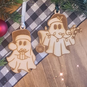 Puede incluir: Dos adornos navideños de madera con forma de fantasmas que llevan gorros de Papá Noel. Un fantasma sostiene una caja de regalo y el otro un bastón de caramelo. Los adornos están sobre un fondo a cuadros blanco y negro.