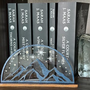 May include: A decorative bookend featuring a mountain range design in blue and silver glitter. The bookend is holding several books by Sarah J. Maas, including "A Court of Silver Flames" and "A Court of Thorns and Roses."