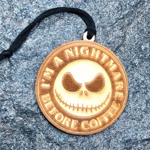 Puede incluir: Un adorno de madera redondo con una cuerda negra para colgar. El adorno presenta una cara de dibujos animados con el texto "I'm a Nightmare Before Coffee".