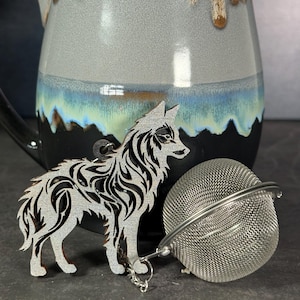 May include: A silver wolf-shaped tea infuser with detailed cut-out patterns, beside a stainless steel mesh tea ball. A ceramic mug with a blue, grey, and brown glaze is in the background.