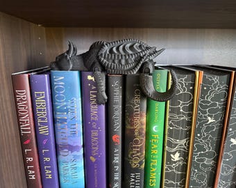 May include: A dark gray dragon-shaped bookend rests atop a row of colorful books. The bookend has a detailed, textured design. The spines of the books are various colors, with titles like "Dragonfall" and "Moon Marked".