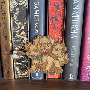 May include: A wooden Cerberus bookmark with three dog heads, holding an axe and a blue staff, rests in front of a row of books. The books have titles like "Anathema" and "Blood of Hercules". The bookmark is brown with detailed engraving.
