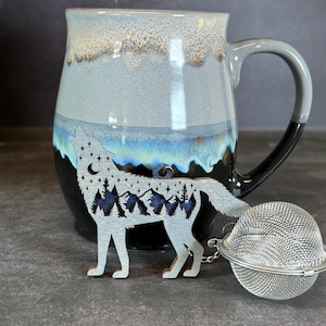 Może przedstawiać: Szary i czarny kubek ceramiczny z wzorem sylwetki wilka. Wilk wyje do księżyca i ma pasmo górskie i drzewa w swojej sylwetce. Kubek jest wyposażony w srebrny metalowy zaparzacz do herbaty.