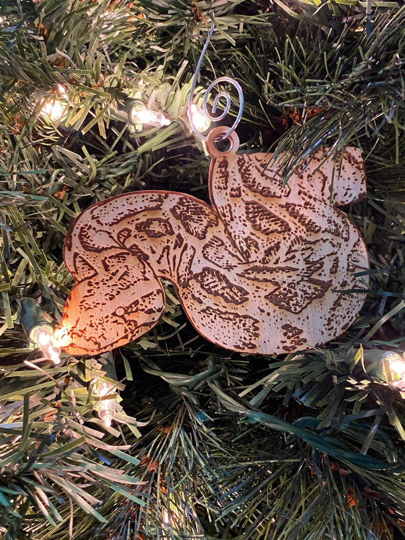 Puede incluir: Adorno navide&ntilde;o de madera con forma de ardilla, con un dise&ntilde;o grabado detallado. El adorno marr&oacute;n claro tiene detalles grabados en marr&oacute;n oscuro y un colgador de alambre plateado. Cuelga de un &aacute;rbol de Navidad verde con luces blancas.