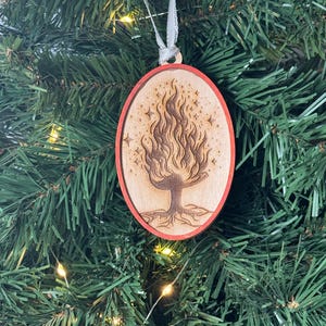 May include: A wooden Christmas ornament shaped like an oval with a red border. The ornament features a laser-cut design of a tree with flames and stars.