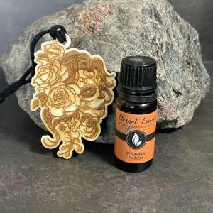 Puede incluir: Un adorno de madera con un diseño de calavera y rosas, colgado de un cordón negro. Una pequeña botella de aceite esencial etiquetada como "Eternal Essence Premium Fragrance Oil Pumpkin Patch" está al lado del adorno.