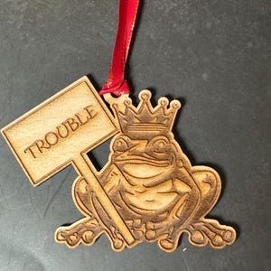 May include: Wooden ornament of a frog wearing a crown and holding a sign that says "Trouble". The ornament is brown and has a red ribbon attached to the top.