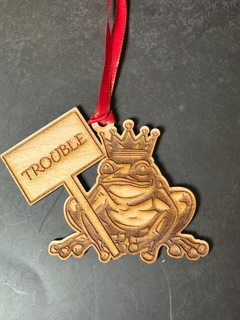 May include: Wooden ornament of a frog wearing a crown and holding a sign that says "Trouble". The ornament is brown and has a red ribbon attached to the top.