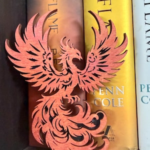 May include: A wooden phoenix bookend in shades of orange and red. The phoenix is intricately cut, with detailed feathers and a stylized design. The bookend is positioned in front of a row of books.