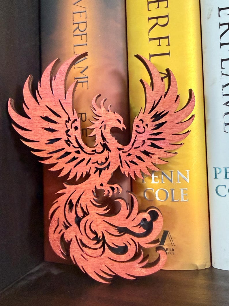 May include: A wooden phoenix bookend in shades of orange and red. The phoenix is intricately cut, with detailed feathers and a stylized design. The bookend is positioned in front of a row of books.
