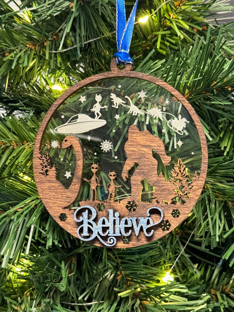 May include: A wooden Christmas ornament with a clear center featuring a Bigfoot, a dinosaur, aliens, and a flying saucer. The ornament is decorated with snowflakes and stars. The text "Believe" is written in silver glitter on the bottom of the ornament.