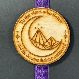 May include: A wooden bookmark with a crescent moon and mountains design. The bookmark has the text "To the stars who listen and the dreams that are answered".
