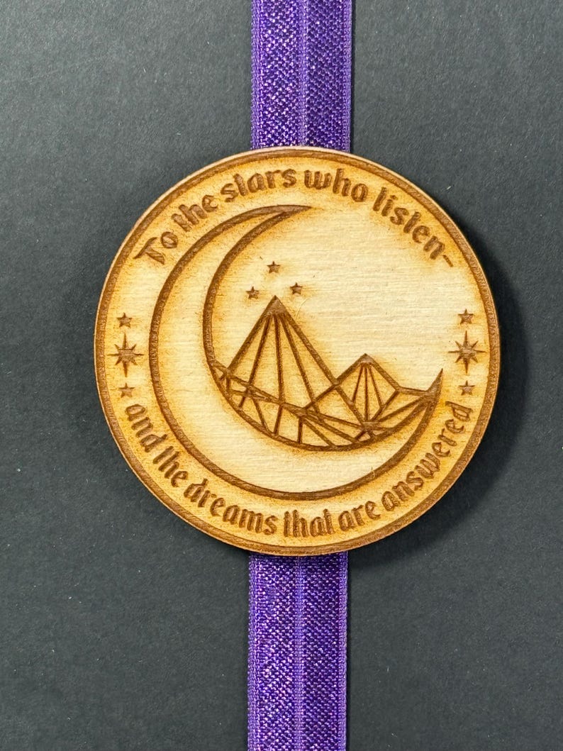 May include: A wooden bookmark with a crescent moon and mountains design. The bookmark has the text "To the stars who listen and the dreams that are answered".
