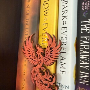 May include: A laser-cut wooden phoenix bookmark in shades of orange and brown rests between three books. The books have titles including "Heat of the Ever", "Glow of the Ever", and "Spark Everflame". The bookmark is a decorative accessory.