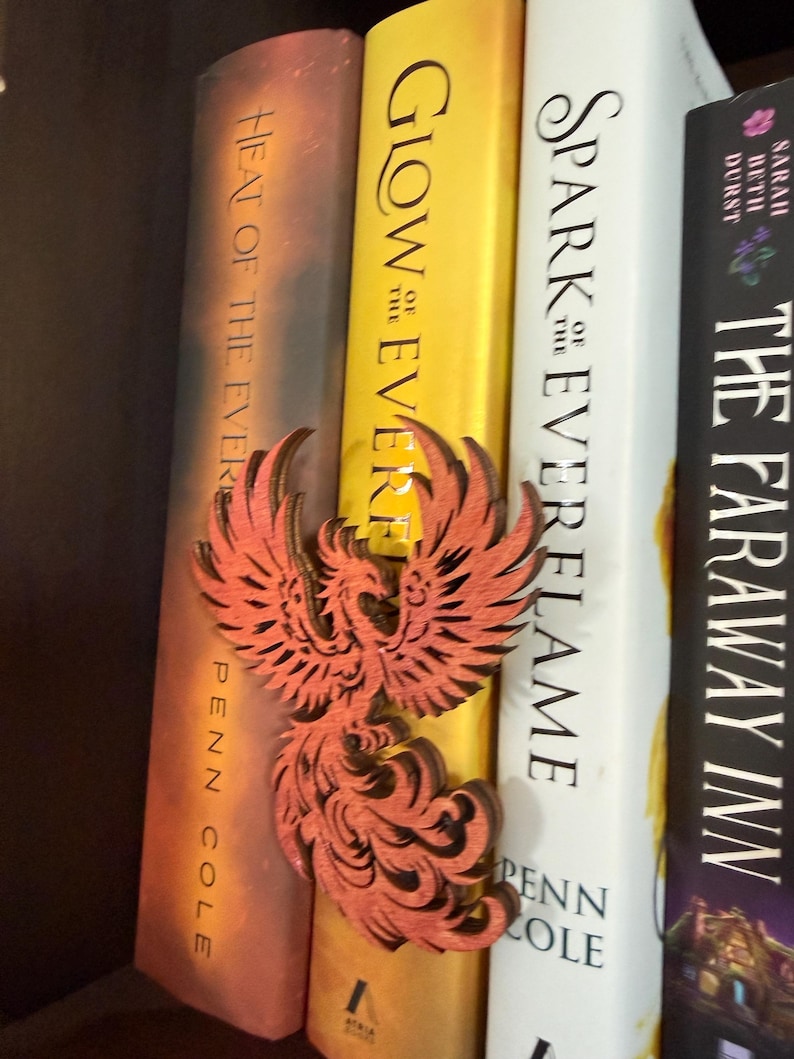 May include: A laser-cut wooden phoenix bookmark in shades of orange and brown rests between three books. The books have titles including "Heat of the Ever", "Glow of the Ever", and "Spark Everflame". The bookmark is a decorative accessory.