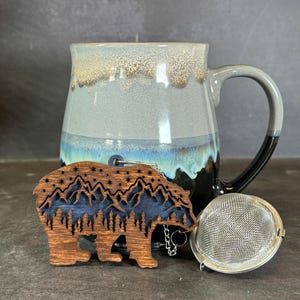 May include: A wooden bear tea infuser with a mountain and star design. The infuser is attached to a metal chain and is next to a silver tea strainer and a blue and black ceramic mug.