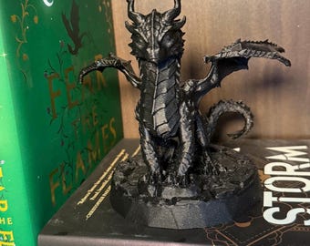 May include: A black 3D printed dragon figurine with wings outstretched, standing on a round base. The dragon has a detailed, textured surface and a fierce expression.