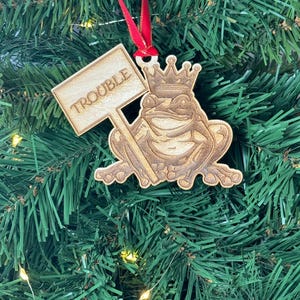 May include: Wooden Christmas ornament featuring a frog wearing a crown and holding a sign that says "Trouble".