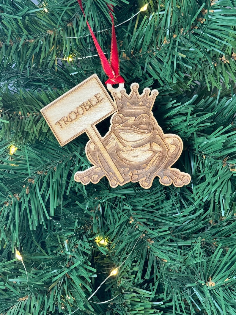 May include: Wooden Christmas ornament featuring a frog wearing a crown and holding a sign that says "Trouble".