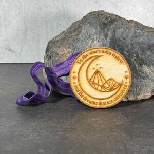 May include: A wooden circle pendant with a crescent moon and stars design. The pendant has the text "To the stars who listen... and the dreams that are answered" engraved on it. The pendant is attached to a purple ribbon.