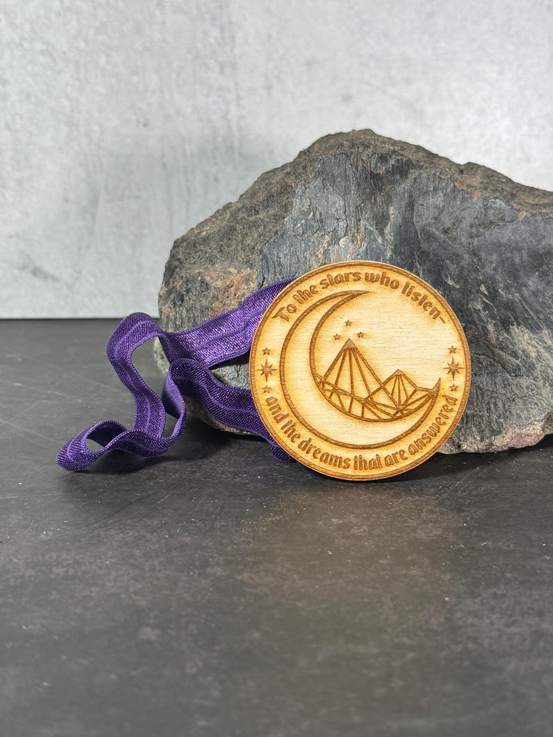 May include: A wooden circle pendant with a crescent moon and stars design. The pendant has the text "To the stars who listen... and the dreams that are answered" engraved on it. The pendant is attached to a purple ribbon.