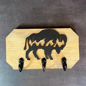 May include: A rectangular wooden wall hook rack featuring a black cutout silhouette of a bison. The bison design contains a negative space landscape of mountains and pine trees. Three black metal hooks are attached to the bottom edge.